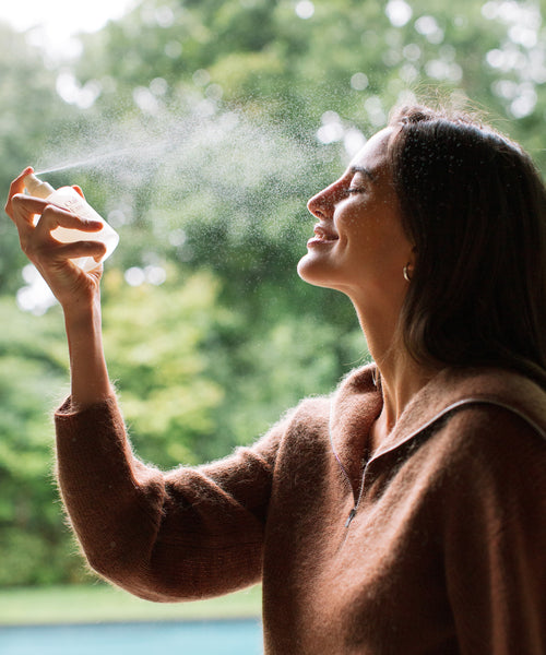 A woman in a brown sweater sprays her face with The Rescue Edit, eyes closed and smiling. Outdoors among green trees and a blurred background, she enjoys a luminous glow.