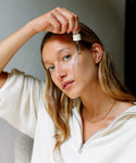 A woman with long blonde hair applies The Rescue Edit serum to her cheek with a dropper, revealing a luminous glow. She wears a white zip-up top and gold chain necklace, looking at the camera in soft, natural light.