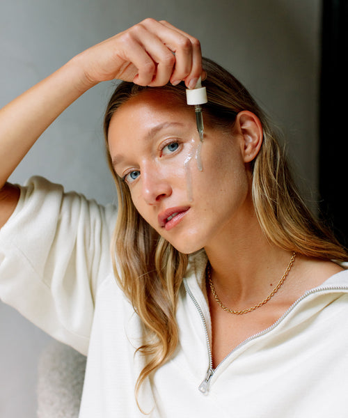 A woman with long blonde hair applies The Rescue Edit serum to her cheek with a dropper, revealing a luminous glow. She wears a white zip-up top and gold chain necklace, looking at the camera in soft, natural light.