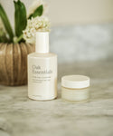 The Double Cleanse Duo from Oak Essentials, featuring a Pure Gel Cleanser and Balancing Balm, is displayed on a marble surface with a vase of white flowers in the background.