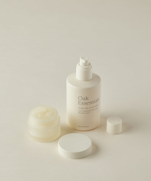Two white containers of Oak Essentials Double Cleanse Duo on a light surface: an open Cleansing Balm jar and a Pure Gel Cleanser pump bottle, with lids nearby—your ideal skincare pair.