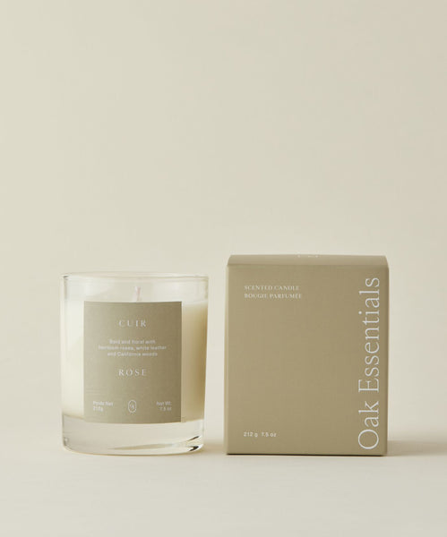 A white Cuir Rose Candle with an heirloom rose scent sits in a clear glass next to a tan Oak Essentials box on a light beige background. Both the candle label and box display minimalist, elegant text.