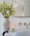A bathroom sink with a marble countertop features the Cuir Rose Candle lit beside a bar of soap on a white towel and a large vase of heirloom roses, with a mirror and wall-mounted faucet above.