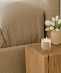 A lit Cuir Rose Candle rests on a wooden side table by a beige bed and white tulips, filling the bedroom with cozy charm and delicate floral-woody fragrance.
