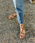 A person in blue jeans stands on a gravel surface wearing Oiled Leather Strappy Sandal.