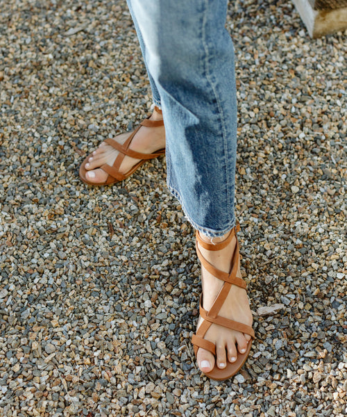 A person in blue jeans stands on a gravel surface wearing Oiled Leather Strappy Sandal.