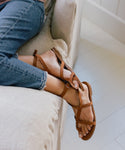 A person in frayed-hem blue jeans relaxes on a beige sofa, showing their feet in brown Oiled Leather Strappy Sandals made in Italy, resting on a light floor.