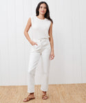 A woman stands indoors on a woven rug, wearing a sleeveless white top, high-waisted white jeans, and Oiled Leather Strappy Sandals with one hand in her pocket, posed against a white paneled wall.