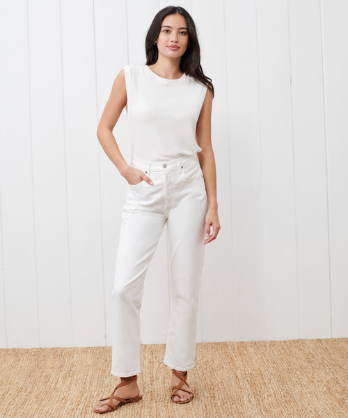 A woman stands indoors on a woven rug, wearing a sleeveless white top, high-waisted white jeans, and Oiled Leather Strappy Sandals with one hand in her pocket, posed against a white paneled wall.
