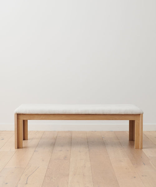 The Otto Bench, featuring a natural linen seat, stands on a wooden floor against a plain white wall.