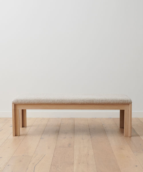 The Otto Bench, crafted from white oak with a light beige wool boucle seat, sits against a plain white wall on a light wood floor.