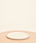 The Picnic Serving Platter from Crow Canyon, in plain white enamel, rests on a light wooden table set against a beige background. The minimal scene features only the platter with no food or other items present.