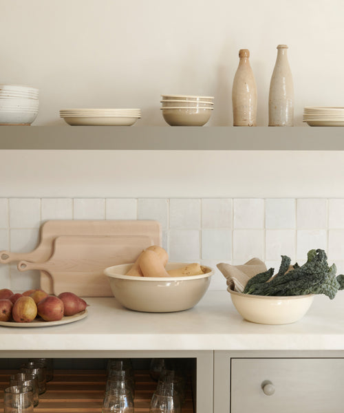 A minimalist kitchen in neutral tones features open shelves with stacked plates, bottles, a Picnic Serving Platter, bowls of squash and potatoes, leafy greens on the counter, and cutting boards resting against a tiled backsplash.