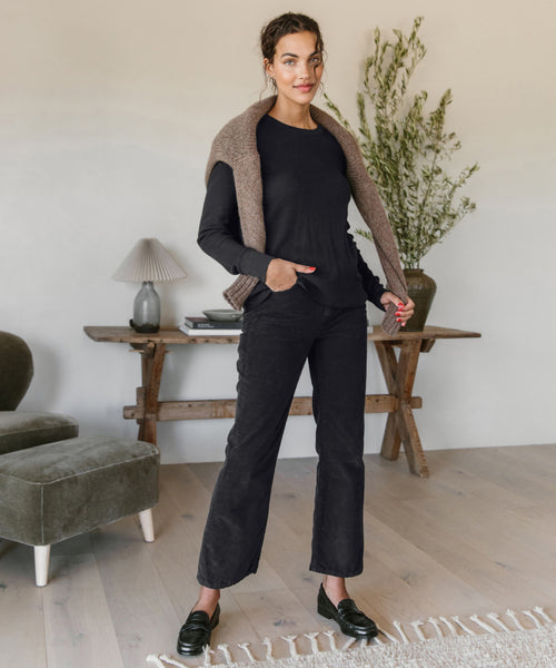 A woman stands indoors with her hands in her pockets, wearing Loose Flare Pant and a washed black long-sleeve top. She has a brown sweater over her shoulders and stands on a light wood floor near a console table and plant.