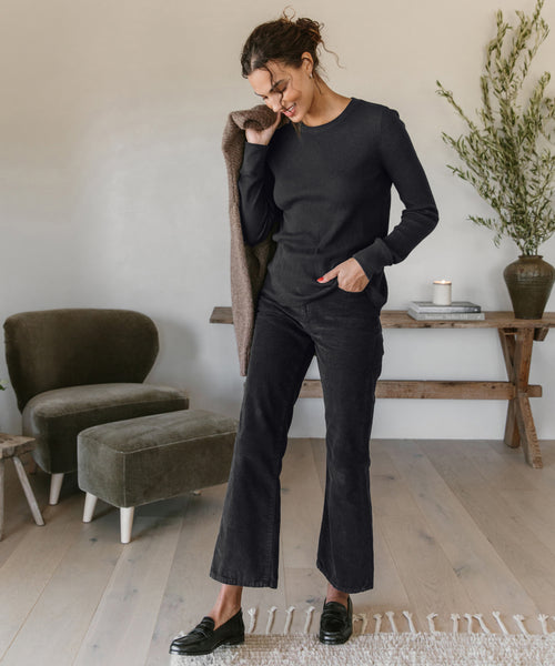 A woman smiles indoors with one hand in her pocket and a coat over her shoulder, wearing a black sweater, loafers, and Loose Flare Pant. The modern room includes stylish furniture, a rug, and a plant on a wooden console table.