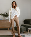A woman stands indoors in a white long-sleeve top, brown slippers, and the Loose Flare Pant in beige. She has a cream sweater draped over her shoulders, with a wooden sideboard, vase of greenery, and dark armchair behind her.
