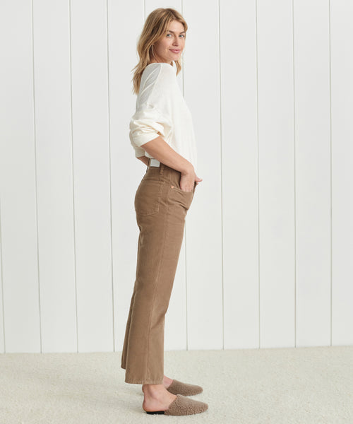 A woman stands in profile against a white paneled wall, wearing a white long-sleeve shirt, brown Loose Flare Pant corduroys, and fuzzy brown slippers, with her hands in her pockets.