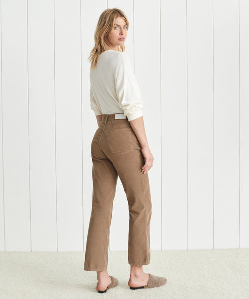 A blonde woman stands indoors, glancing over her shoulder. She wears a white long-sleeve top, Loose Flare Pant in tan corduroy, and fuzzy slippers, set against a white paneled wall and carpeted floor.