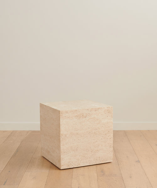The Sur Side Table, a beige, stone-like cube inspired by solid travertine, sits on a light wooden floor against an off-white wall in a minimalist room.