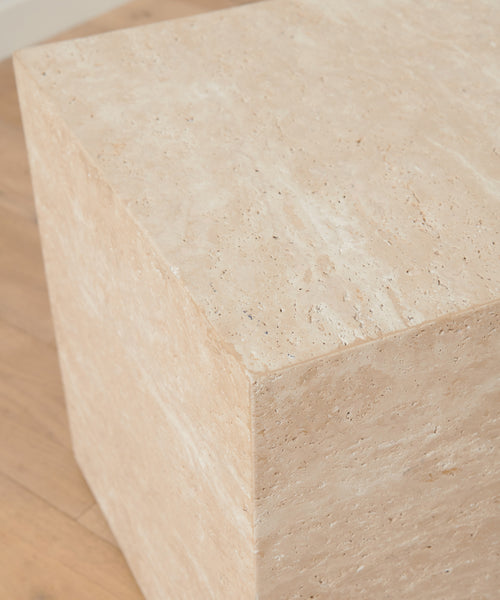The Sur Side Table, a large beige travertine block with clean edges, sits on a light wooden floor and is illuminated by soft natural light.