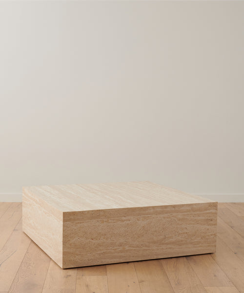 The Sur Square Coffee Table, a minimalist rectangular piece made of light-colored travertine stone, stands on a wooden floor against an off-white wall in a modern interior.