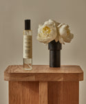 A bottle of Topanga Room Spray, crafted in Los Angeles with nature-inspired scents, sits beside a small black vase holding white flowers on a wooden table against a neutral backdrop.