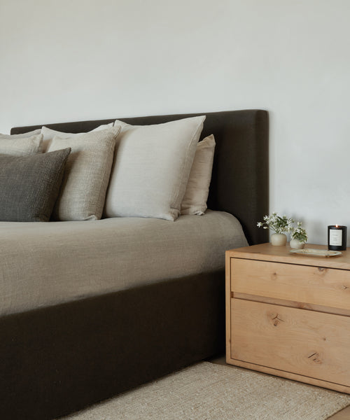 A neatly made Pacific Bed with an upholstered headboard and Belgian Libeco linen pillows sits beside a light wooden nightstand holding small vases of white flowers and a lit candle, set against a plain white wall.