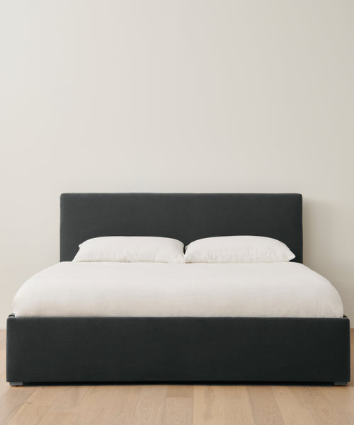 The Pacific Bed features a dark upholstered headboard and frame, topped with white bedding and two white pillows. Positioned on a light wood floor against an off-white wall, it adds timeless charm to the minimalist space.