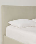 A close-up of a neatly made bed featuring a light beige upholstered headboard in Fabric Swatch Stone Linen, with white pillow and bedding against a plain white wall.