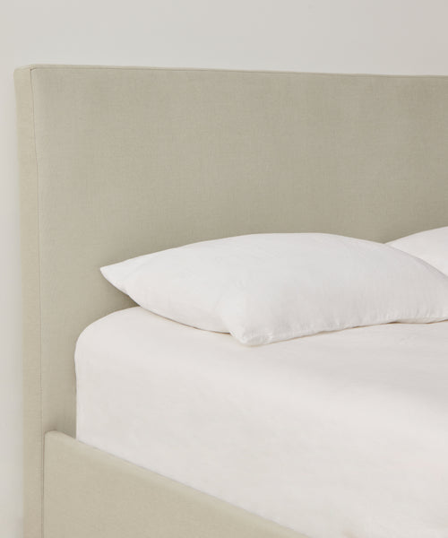 A close-up of a neatly made bed featuring a light beige upholstered headboard in Fabric Swatch Stone Linen, with white pillow and bedding against a plain white wall.