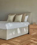 A minimalist bedroom featuring the Pacific Bed with a light beige upholstered headboard, white bedding, neutral pillows, a wooden nightstand, and a natural fiber rug on the floor against a light gray wall.