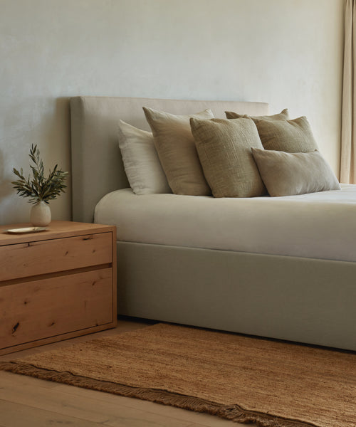 The Pacific Bed, featuring an upholstered headboard and Belgian linen bedding, stands next to a wooden nightstand with a vase of greenery. A woven rug is placed beside the bed on the wooden floor.