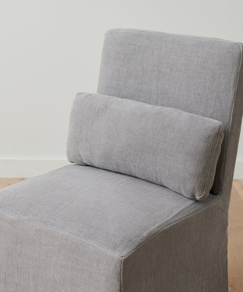 The Sunset Dining Chair Slipcover, in light gray ash linen, covers a modern armless chair and includes a rectangular matching lumbar pillow, shown against a white wall and wooden floor.