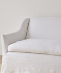 A close-up of the Miramar Sofa Slipcover in white linen upholstery against a plain light wall, highlighting its armrest, backrest, and seat cushion. The slipcover features a simple, minimalist design.