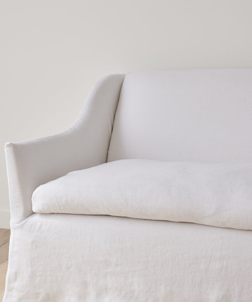 A close-up of the Miramar Sofa Slipcover in white linen upholstery against a plain light wall, highlighting its armrest, backrest, and seat cushion. The slipcover features a simple, minimalist design.