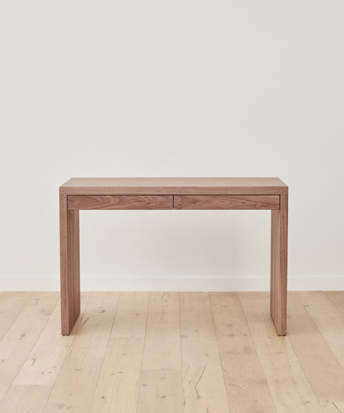 The Newport Desk, featuring two understated drawers, stands against a plain white wall on a light wood floor, bringing a simple yet timeless vintage-inspired look to your space.