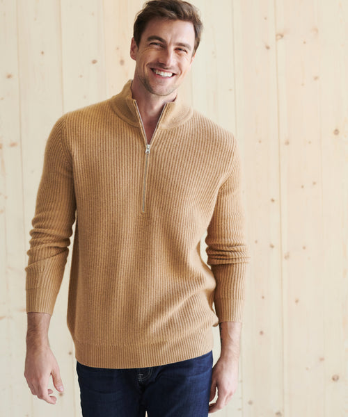 A smiling man in a tan Mens Wool Half Zip and dark jeans stands before a light wooden wall.