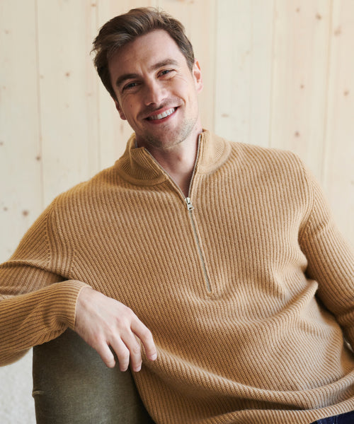 A smiling man with short brown hair wears the Mens Wool Half Zip sweater and sits comfortably on a green chair, set against a light wood-paneled background.