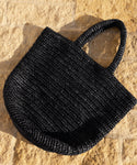 The Woven Raffia Tote with two handles in black is displayed on a textured, light tan stone surface under sunlight.