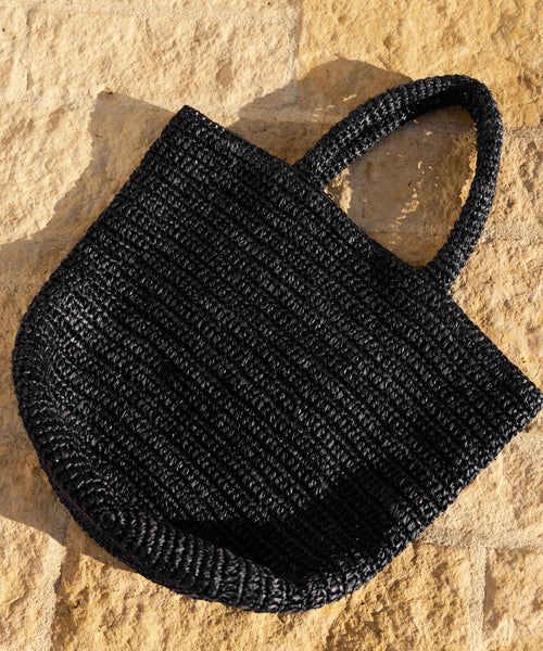 The Woven Raffia Tote with two handles in black is displayed on a textured, light tan stone surface under sunlight.