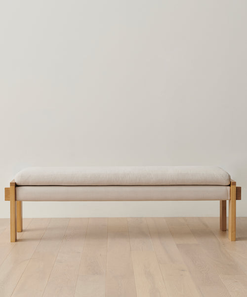The Ynez Bench, a minimalist luxury entryway bench in White Oak with a light beige Belgian Libeco linen cushion, sits on a light wood floor against an off-white wall, radiating timeless elegance.