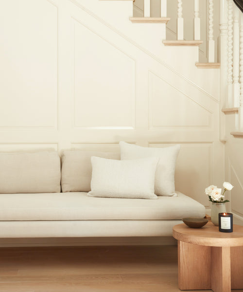 The Ynez Daybed in beige Belgian linen with two cushions sits before a white paneled staircase. A wooden side table nearby displays a vase of white flowers, candle, and bowl in the bright, minimalist living space.