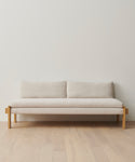 The Ynez Daybed, featuring a minimalist Belgian linen design with wooden legs and frame, sits against a plain white wall on light wood flooring. It has two back cushions and a simple, modern look.