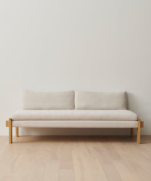 The Ynez Daybed, featuring a minimalist Belgian linen design with wooden legs and frame, sits against a plain white wall on light wood flooring. It has two back cushions and a simple, modern look.