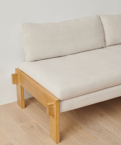 The Ynez Daybed features a minimalist design with a light linen-cushioned seat and backrest, set on a natural wooden frame and legs. Its shown placed on a light wood floor against a plain white wall.
